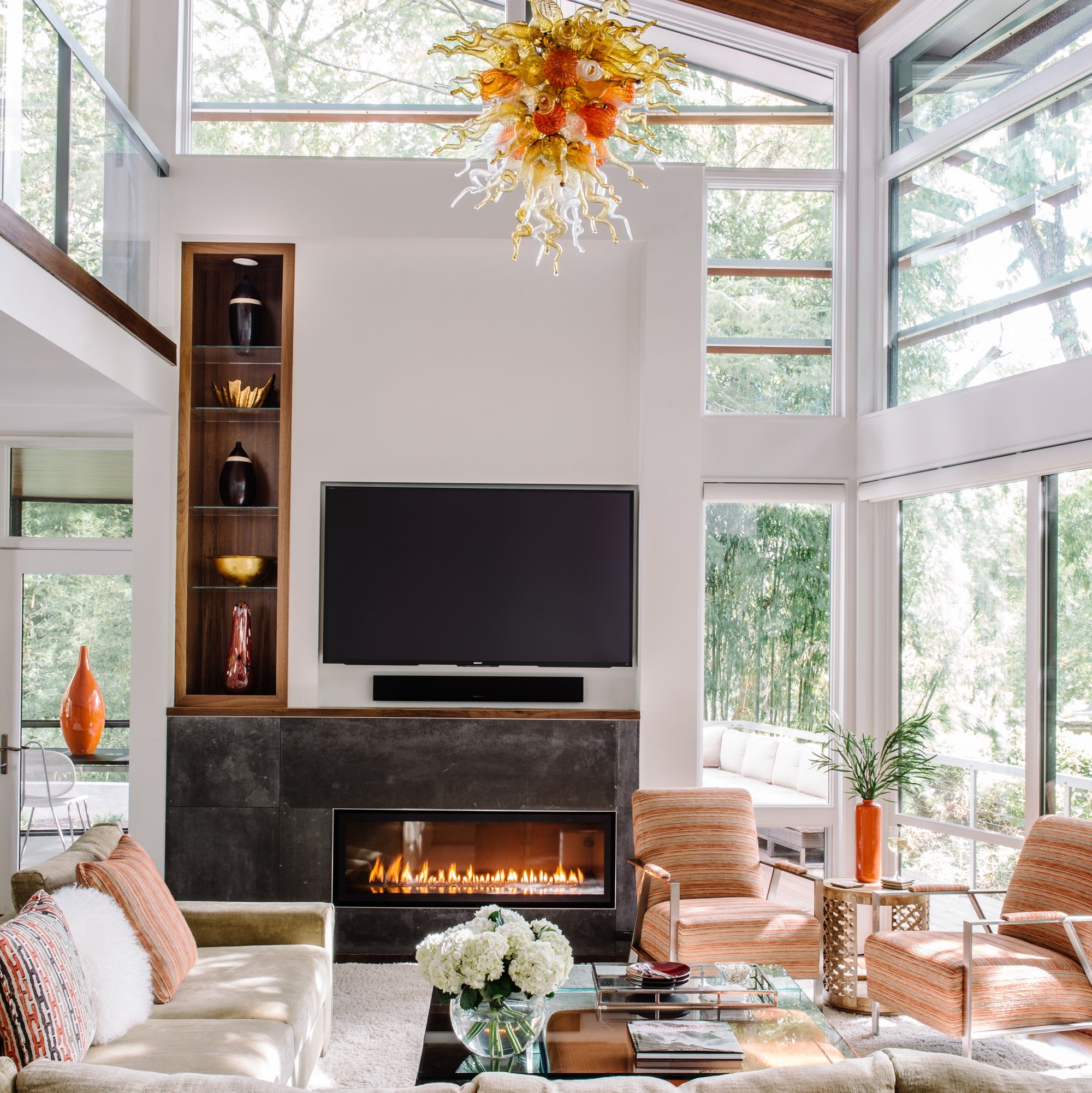Family room with mid-century modern style featuring art glass chandelier, slate fireplace, and neutral and orange furnishings. Jamie Merida Interiors.